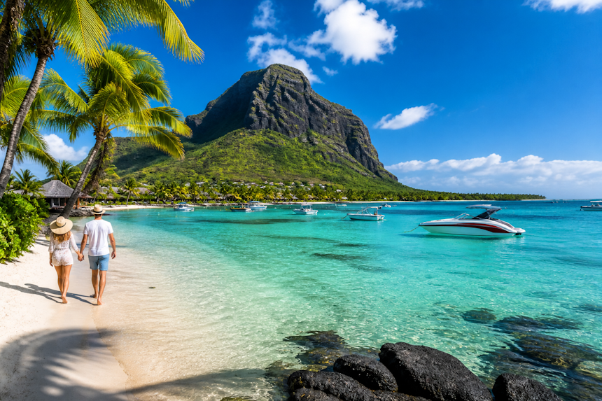 Mauritius tropical beach with turquoise lagoon, white sand, palm trees, luxury resort, and a couple of tourists walking along the shoreline with boats in clear water and a mountain backdrop.